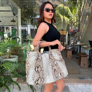 May include: A large tote bag featuring a textured snakeskin pattern in shades of beige, brown, and cream. The bag has two rounded top handles and a spacious rectangular body. It is constructed with a distinct scale-like surface texture throughout.