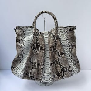 May include: A large tote bag featuring a textured snakeskin pattern in shades of gray, brown, and white. The bag has a rounded top handle, a pleated design that creates a soft, gathered silhouette, and silver-toned hardware attachments on the sides.