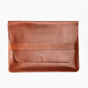 May include: A brown leather laptop sleeve with a flap closure and a securing strap. The sleeve has a minimalist design with a subtle logo on the front. The leather has a natural, textured appearance.