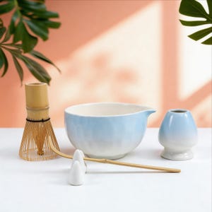 May include: A matcha tea set featuring a bamboo whisk, a light blue ceramic bowl, a small vase, and two white ceramic rests. The set is arranged on a white surface with a peach-colored background and green foliage.