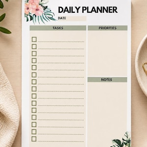 May include: A white daily planner with floral accents. The planner features sections for tasks, priorities, and notes, with a space for the date. The words "Daily Planner" are printed at the top in black.