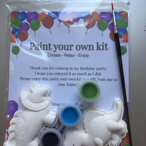 Könnte beinhalten: Ein Mal-dich-selbst-Set in einer transparenten Plastiktüte. Das Set enthält zwei weiße Dinosaurierfiguren, drei kleine Farbtöpfe in Blau, Grün und Schwarz sowie einen Pinsel. Die Verpackung trägt den Text "Paint your own kit".