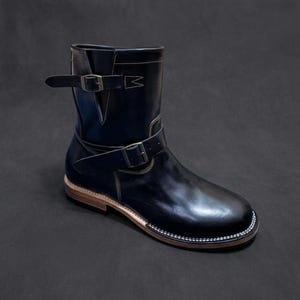 Könnte beinhalten: Ein schwarzer Leder-Ingenieurstiefel mit abgerundeter Zehenpartie, einem gestapelten Holzabsatz und einem hellen Rahmen mit weißen Nähten. Der Stiefel verfügt über zwei verstellbare Riemen mit Metallschnallen, einen am Knöchel und einen am Schaft.