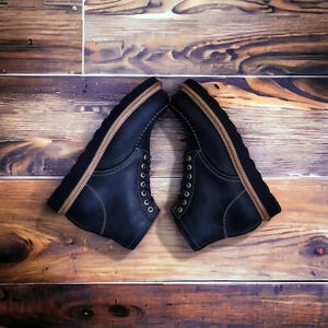 以下が含まれることがあります： モックトゥデザインの黒いレザーブーツ一足。白いコントラストステッチ、真鍮色の金属製アイレット、ナチュラルなタンカラーのレザーウェルトが付いた厚手の黒いラグソールが特徴です。ブーツは向かい合わせに配置されています。