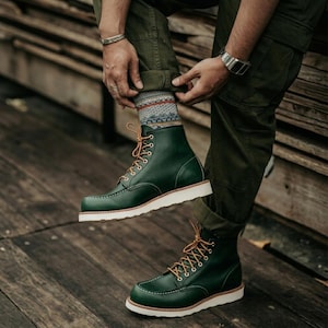 Puede incluir: Un par de botas de cuero verde oscuro con puntera estilo mocasín, cordones de color marrón claro, ojales metálicos plateados y una suela gruesa de goma blanca. Las botas se llevan con calcetines estampados con rayas horizontales en azul, naranja y blanco.