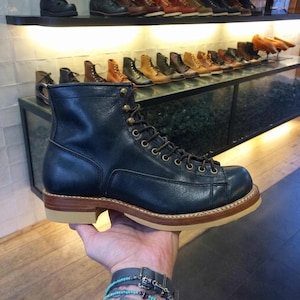 Puede incluir: Una bota de cuero azul marino oscuro con cordones, punta redondeada, ojales metálicos en tono latón, construcción de vira visible y una suela gruesa de goma color canela claro. La bota presenta un cuello estructurado y acabado liso.