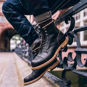 Könnte beinhalten: Ein schwarzer Schnürstiefel aus Leder mit einer Zehenkappe, die dekorative Brogue-Perforationen aufweist. Der Stiefel hat eine hellbraune Ledermittelsohle und einen Schichtabsatz, schwarze Schnürsenkel mit silberfarbenen Ösen und einen beigen Lederbesatz.