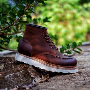 Könnte beinhalten: Ein brauner Lederstiefel mit einer genähten Mokassin-Spitze, dunkelbraunen Schnürsenkeln, Metallösen und einer dicken weißen Keilsohle aus Gummi. Der Stiefel weist sichtbare Nähte entlang des Rahmenbesatzes und eine matte Lederoberfläche auf.