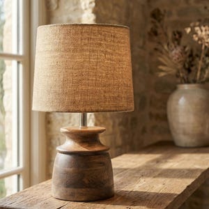 Puede incluir: Una lámpara de mesa de madera con una pantalla de arpillera. La lámpara tiene una base de madera tallada de color marrón oscuro y un vástago de metal plateado. La pantalla es de color marrón claro. La lámpara está sobre una mesa de madera rústica.