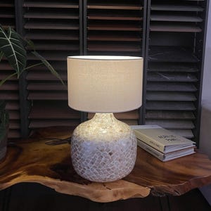 Può includere: Una lampada da tavolo decorativa con paralume beige e base strutturata bianco sporco. La lampada è su un tavolo in legno scuro, accanto a dei libri. La base della lampada presenta un motivo a mosaico.