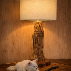 May include: A wooden table lamp with a beige lampshade. The lamp has a textured, natural wood base. A white cat with a yellow collar is lying on a wooden table.