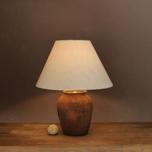 Puede incluir: Una lámpara de mesa decorativa con una pantalla beige y una base marrón texturizada. La base tiene un diseño grabado. La lámpara está sobre una superficie de madera. Una pequeña bola decorativa está al lado de la lámpara.