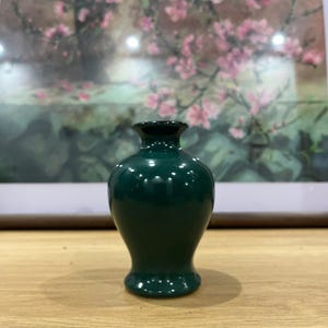 Puede incluir: Un pequeño jarrón de cerámica verde oscuro con un acabado brillante. El jarrón tiene un cuerpo redondeado, una base acampanada y un cuello estrecho con un borde negro. El fondo presenta una pintura de flores rosas.