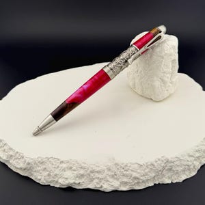 May include: A handcrafted pen with a vibrant pink and brown barrel, accented with silver-colored metal detailing. The pen rests on a white, textured surface against a dark background. The pen's design features intricate patterns.