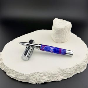 May include: A silver fountain pen with a cap, featuring a vibrant barrel design of blue and purple swirls. The pen is displayed on a textured white surface, with a small white decorative object in the background.