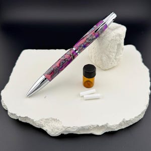 May include: A handcrafted pen with a vibrant purple, pink, and silver marbled design. The pen has a silver tip and clip. Also included are a small amber bottle with a black cap and two white refills. The pen rests on a white, textured surface.