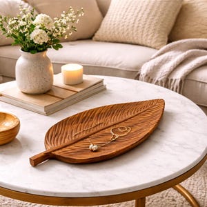 Op de afbeelding: Een houten blad-vormige dienblad met gesneden details bevat delicate gouden kettingen en een ring. De dienblad staat op een wit marmeren tafel met een gouden basis. Een vaas met witte bloemen, een kaars en boeken staan ook op de tafel.