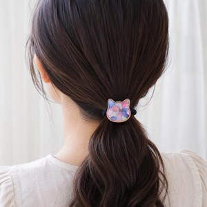 Könnte beinhalten: Ein Haargummi mit einem katzenförmigen Anhänger, der einen goldfarbenen Metallrand und eine mosaikartige Einlage aus bunten, durchscheinenden Teilen in Rosa, Blau, Violett und Weiß auf einem dunklen elastischen Haarband aufweist.