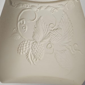 May include: Off-white ceramic bottle with a detailed embossed grape and vine design. The relief carving features clusters of grapes, leaves, and curling vines, creating a textured surface. The bottle has a slightly curved shape.