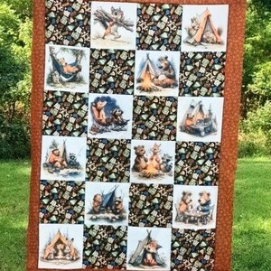 May include: A rectangular patchwork quilt featuring alternating squares of camping-themed illustrations and a dark patterned fabric. The quilt is bordered by a solid burnt orange fabric and displays various woodland animals in outdoor settings.