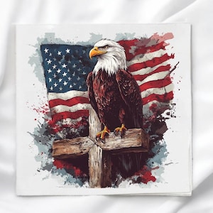 May include: A square print featuring a bald eagle with a white head and brown body perched on a wooden cross. Behind the eagle is a stylized American flag with red, white, and blue colors, accented by dark paint splatter effects on a white background.