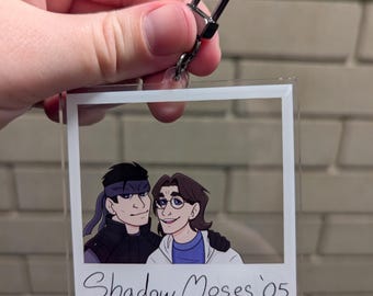 May include: Clear acrylic keychain featuring a cartoon image of two people in a polaroid frame. The image has the text "Shadow Moses '05" in a handwritten style. The keychain has a silver-colored clasp.