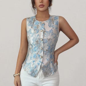 May include: A sleeveless vest featuring a light blue and white floral jacquard pattern with a subtle metallic sheen. The garment has a round neckline and a front button closure with five round, white, pearl-like buttons arranged vertically.