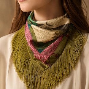 May include: A square silk scarf featuring a detailed print of white birds and green foliage, accented by a wide pink border. The scarf is finished with long, olive green fringe trim along the edges, worn folded in a triangular shape around the neck.