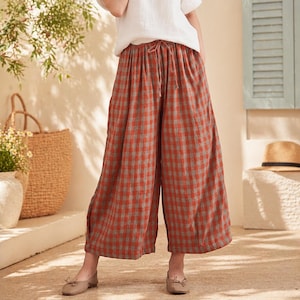 May include: Wide-leg, rust-colored plaid pants with a drawstring waist. The pants have a relaxed fit and are paired with neutral-toned shoes. The plaid pattern features a mix of rust and gray tones, creating a casual, comfortable look.