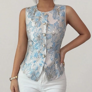 May include: A sleeveless waistcoat featuring a light blue and white floral pattern with a subtle metallic sheen. The garment has a round neckline and a front button closure with five round, white, pearl-like buttons. The fabric has a textured finish.