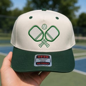 K&ouml;nnte beinhalten: Eine cremefarbene 5-Panel-Kappe mit dunkelgr&uuml;nem Schirm und gr&uuml;n gestickten Pickleball-Schl&auml;gern sowie einem Ball auf der Vorderseite. Ein Aufkleber auf dem Schirm zeigt OTTO WORLD FAMOUS FOAM FRONT OSFM MID PROFILE MAXIMUM COMFORT.