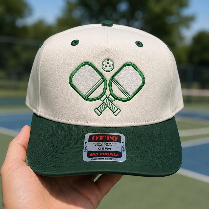 K&ouml;nnte beinhalten: Eine cremefarbene 5-Panel-Kappe mit dunkelgr&uuml;nem Schirm und gr&uuml;n gestickten Pickleball-Schl&auml;gern sowie einem Ball auf der Vorderseite. Ein Aufkleber auf dem Schirm zeigt OTTO WORLD FAMOUS FOAM FRONT OSFM MID PROFILE MAXIMUM COMFORT.