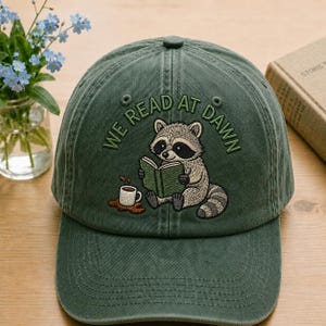 May include: A forest green baseball cap featuring an embroidered graphic of a raccoon reading a green book with a spilled cup of coffee. The text "WE READ AT DAWN" is arched above the raccoon in dark green thread. The cap has a structured crown.