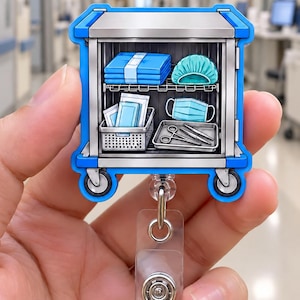 May include: A medical supply cart badge reel featuring a blue and silver frame. The cart has two shelves containing folded blue linens, a teal surgical cap, a light blue face mask, a white basket with medical packets, and a metal tray with tools.