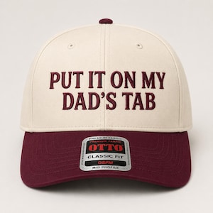 May include: A cream-colored baseball cap with a maroon brim and maroon embroidered text that reads "PUT IT ON MY DAD'S TAB". The cap features a central button on top and a sticker on the brim that reads "OTTO CLASSIC FIT OSFM MID PROFILE".