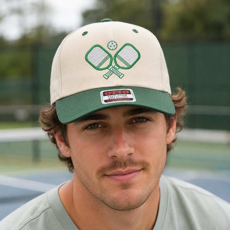 K&ouml;nnte beinhalten: Eine cremefarbene Baseballkappe mit dunkelgr&uuml;nem Schirm und einem gestickten Motiv aus zwei gekreuzten Pickleball-Schl&auml;gern und einem Ball. Ein rechteckiger Aufkleber auf dem Schirm zeigt den Text OTTO FIVE STAR Mid Profile SNAPBACK.