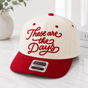 May include: A cream-colored baseball cap with a red brim and top button. The phrase "These are the Days" is embroidered in red script across the front. The cap has a classic fit and a mid profile.