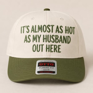 May include: A cream-colored baseball cap with an olive green brim and matching embroidered text that reads: IT'S ALMOST AS HOT AS MY HUSBAND OUT HERE. The cap features a rectangular sticker on the brim with the text THE AUTHENTIC OTTO CLASSIC FIT OSFM.