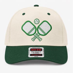 K&ouml;nnte beinhalten: Eine cremefarbene Baseballkappe mit f&uuml;nf Paneelen, dunkelgr&uuml;nem Schirm und passenden gr&uuml;nen &Ouml;sen. Auf der Vorderseite befindet sich eine Stickerei von zwei gekreuzten wei&szlig;en Pickleball-Schl&auml;gern mit gr&uuml;nen Umrandungen und einem Ball.