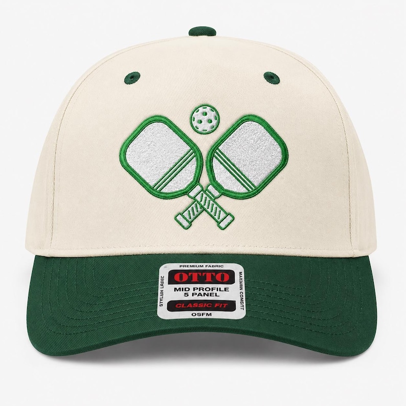 K&ouml;nnte beinhalten: Eine cremefarbene Baseballkappe mit f&uuml;nf Paneelen, dunkelgr&uuml;nem Schirm und passenden gr&uuml;nen &Ouml;sen. Auf der Vorderseite befindet sich eine Stickerei von zwei gekreuzten wei&szlig;en Pickleball-Schl&auml;gern mit gr&uuml;nen Umrandungen und einem Ball.