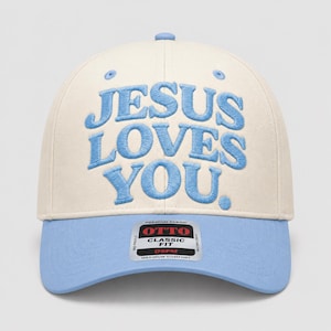 May include: A cream-colored baseball cap with light blue embroidery on the front that reads JESUS LOVES YOU. The cap features a light blue brim and a rectangular sticker on the brim with the text OTTO CLASSIC FIT OSFM MAXIMUM COMFORT.