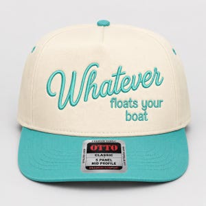 May include: A cream-colored five-panel cap with a teal brim and matching teal button on top. The front features embroidered teal text that reads "Whatever floats your boat" in a mix of cursive and block lettering. A sticker is on the brim.