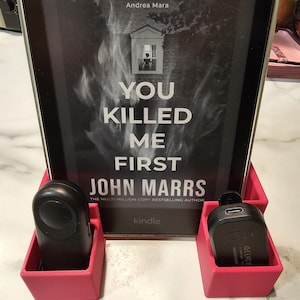May include: A black Kindle e-reader displaying the book cover for "You Killed Me First" by John Marrs, held in a pink stand with two black accessories. The cover features white text and a graphic of a house.