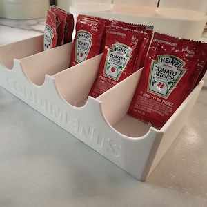 May include: A white plastic condiment organizer with four compartments, each holding red Heinz Tomato Ketchup packets. The word "CONDIMENTS" is embossed on the front of the organizer.