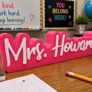 May include: A bright pink, three-dimensional desk sign featuring the name "Mrs. Howard" in raised white script lettering. The sign has a textured, matte finish and is positioned on a wooden surface, displaying a bold, clear font style.