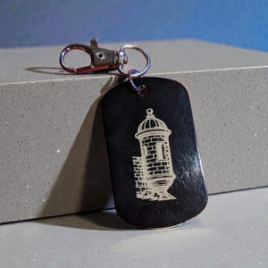 May include: Black rectangular keychain with a silver clasp. The keychain features a detailed engraving of a stone tower with a domed roof. The tower is in a light color, contrasting against the black background. The keychain is made of a durable material.