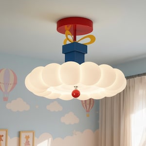 May include: A whimsical ceiling light fixture in a gift box design. The fixture features a red top, blue box, and a cloud-shaped light with a red ornament. The light is designed for a child's room, with a sky blue wall and hot air balloon decals.