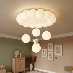 May include: A whimsical ceiling light fixture featuring a cloud-shaped design with multiple hanging globe lights. A teddy bear decorates one of the globe lights. The fixture is white and designed for a nursery or children's room.