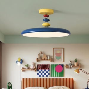 Może przedstawiać: Nowoczesna, kolorowa lampa sufitowa. Lampa ma duży, niebieski, okrągły klosz z białym światłem. Nad kloszem znajdują się ułożone w stos elementy w kształcie dysków w kolorze żółtym, czerwonym i niebieskim. Idealna do pokoju dziecka.