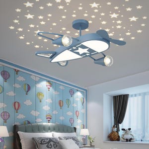 May include: A blue airplane-shaped ceiling light fixture with a white star design on the underside. The light projects star shapes onto the ceiling. The room has a blue and white wallpaper with hot air balloons.